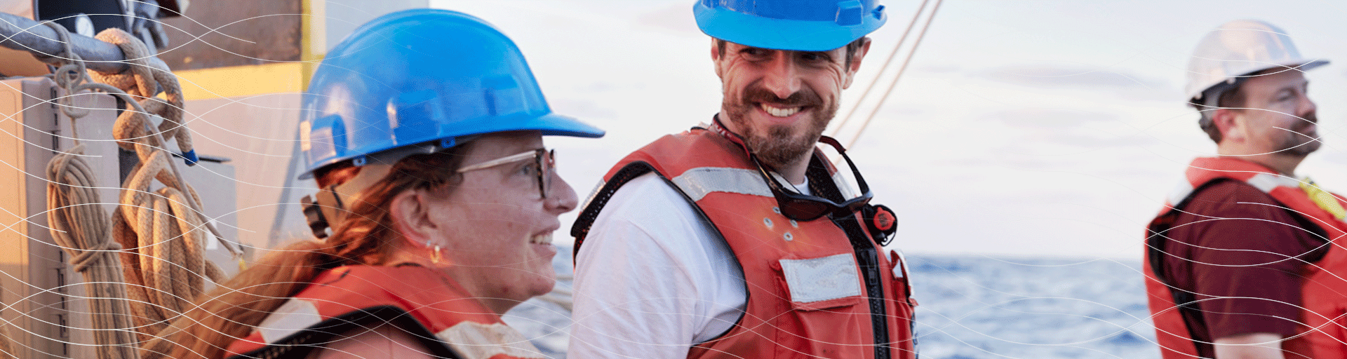Three adults wearing hard hats and safety vests standing on a ship at sunrise or sunset with the ocean visible in the background